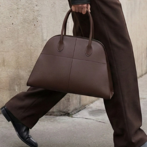 Person walking on a sidewalk wearing a black leather jacket and brown pants, holding a brown handbag.