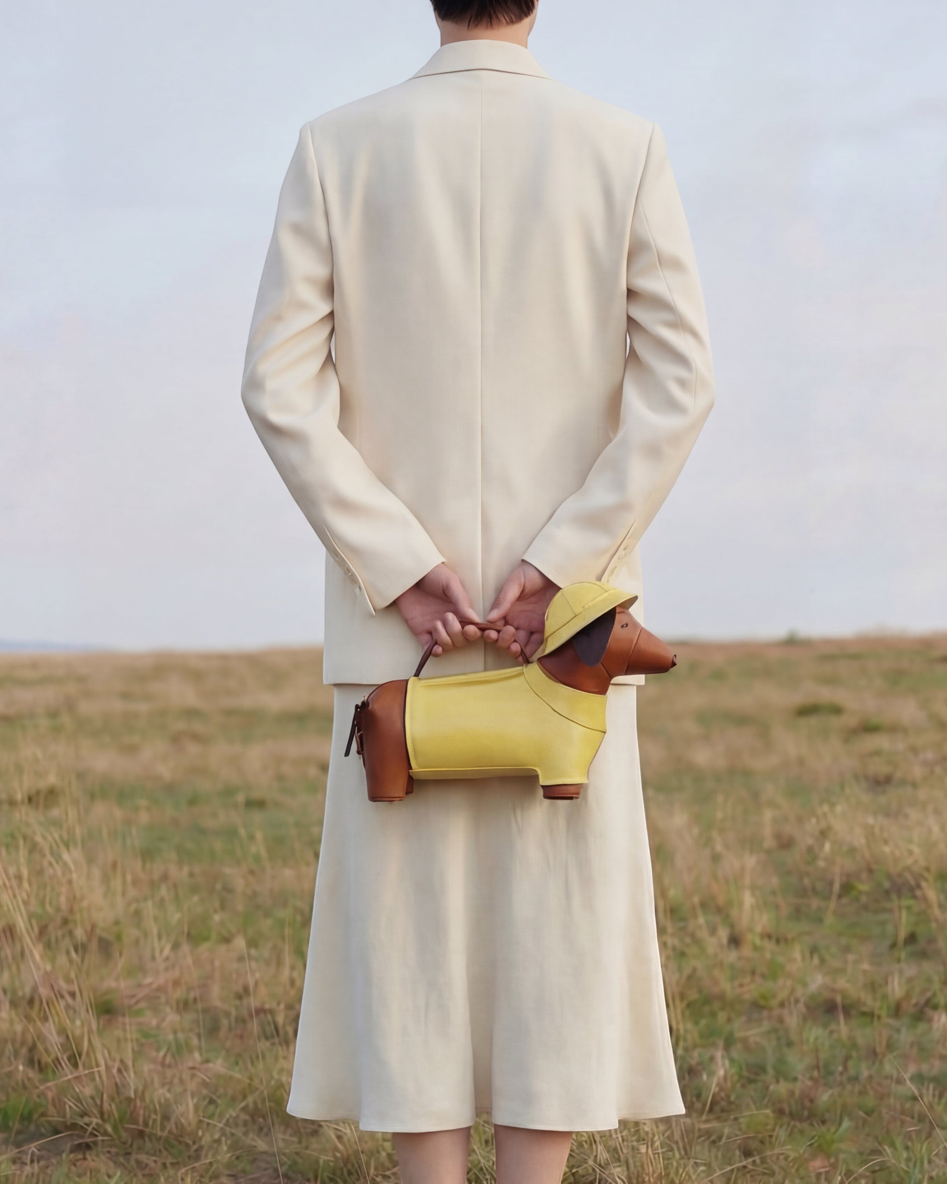 Pine Drew India Dachshund Safari Bag in yellow and brown vegan leather held by a person in a cream suit outdoors.