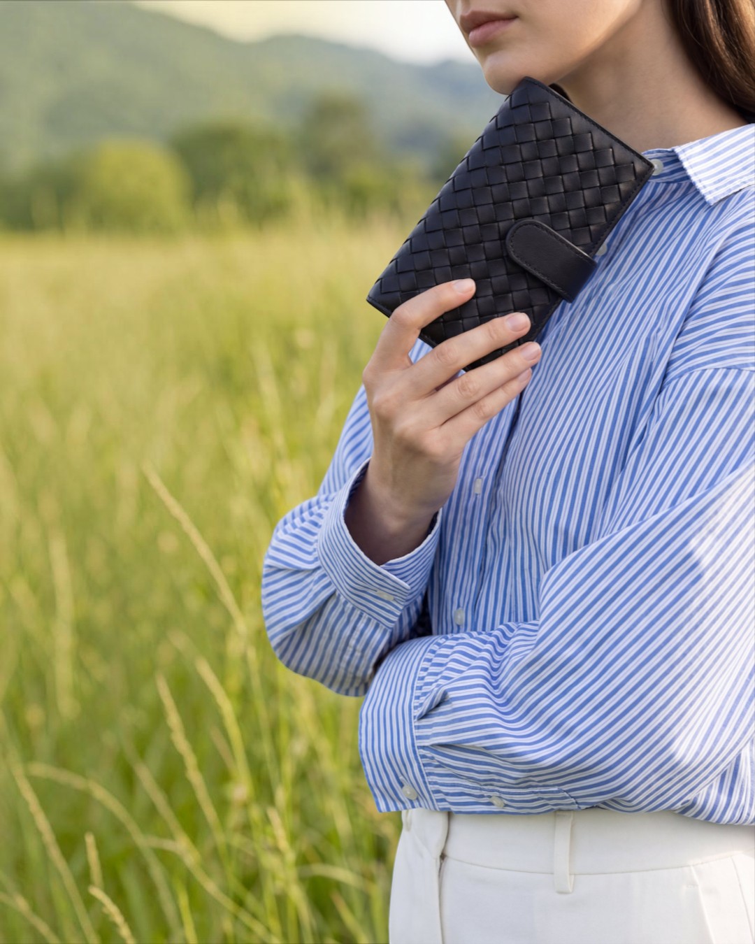 Pine Drew India Caden Interlace long wallet in black woven leather held by a person in a striped shirt in a field.