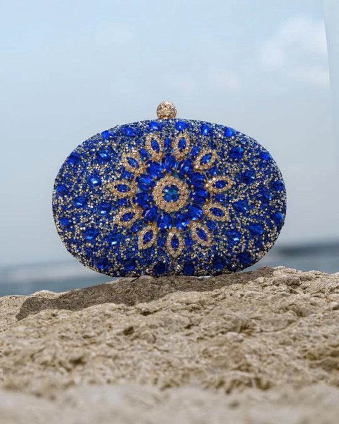 Pine Drew India Colette clutch bag in royal blue with intricate diamond-like crystal embellishments on a rocky beach.